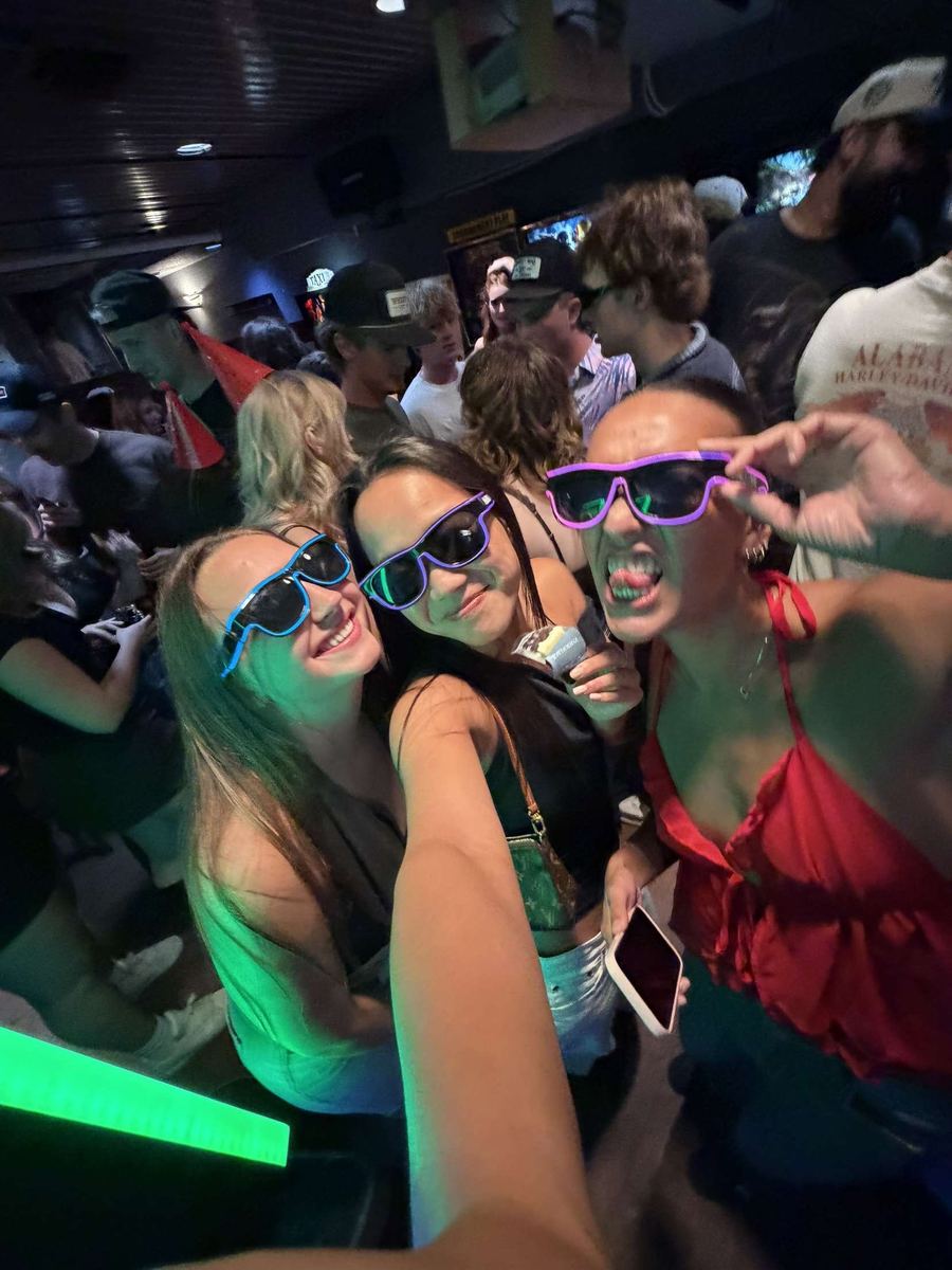 Three friends in a packed bar wearing blue, purple and pink Sunglasses At Night LED glasses