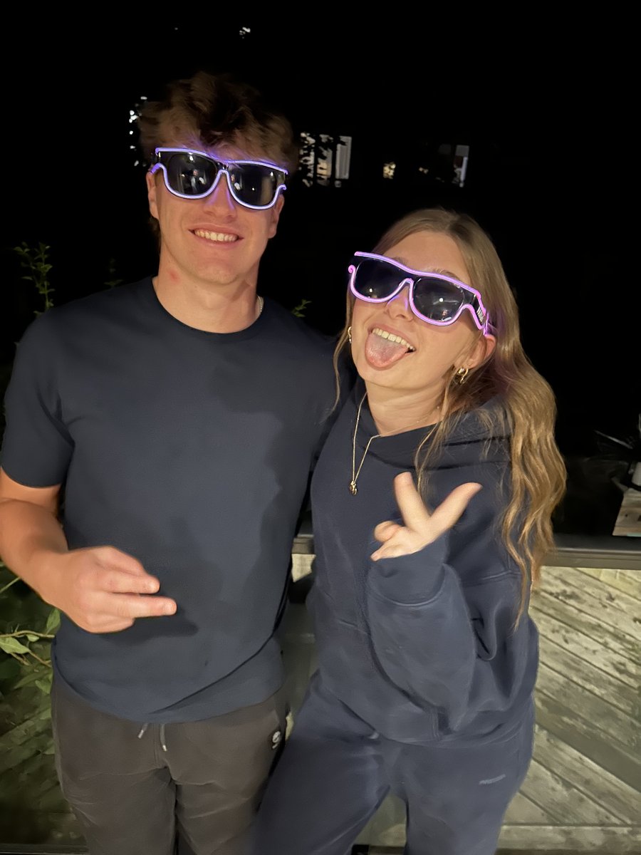 Two friends wearing purple Sunglasses At Night glasses, one making a peace sign