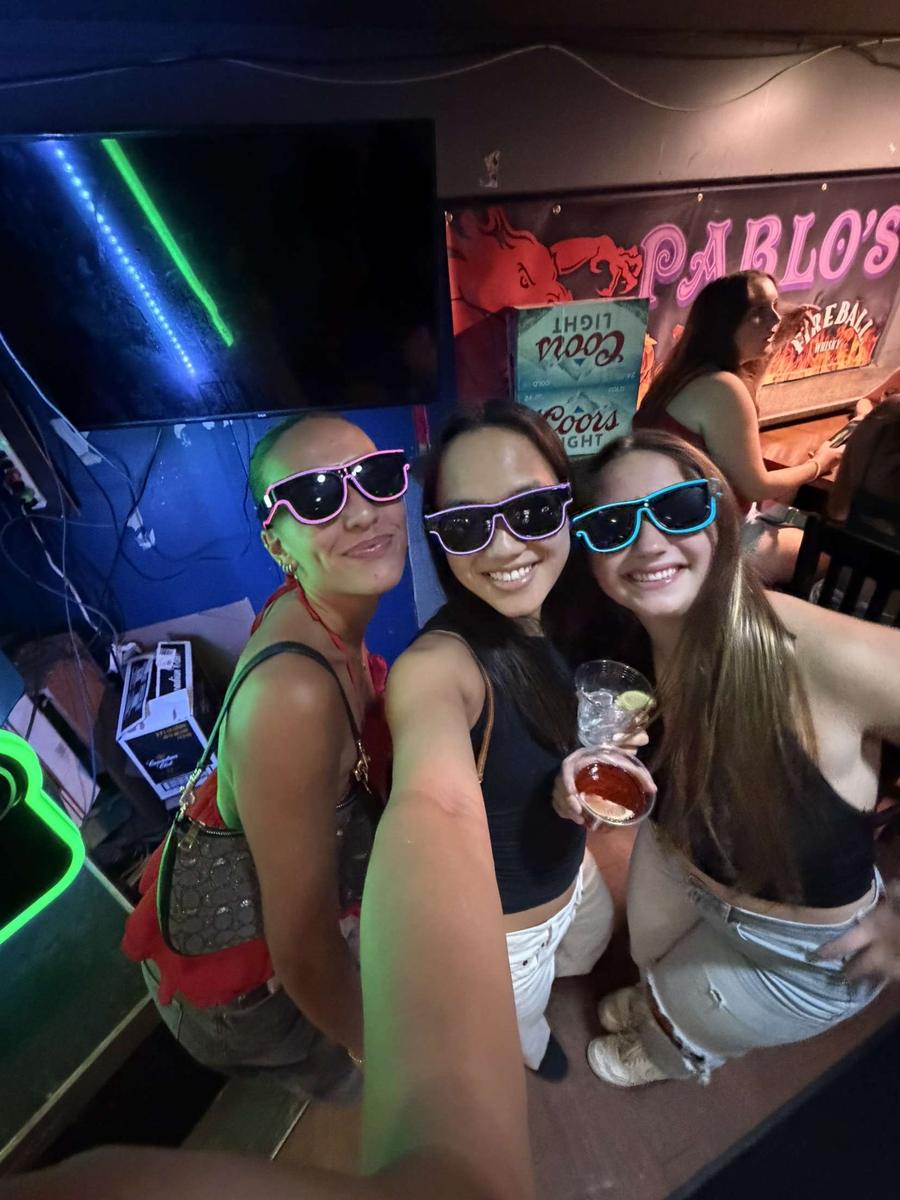 Three friends at a bar wearing pink, purple and blue Sunglasses At Night LED glasses